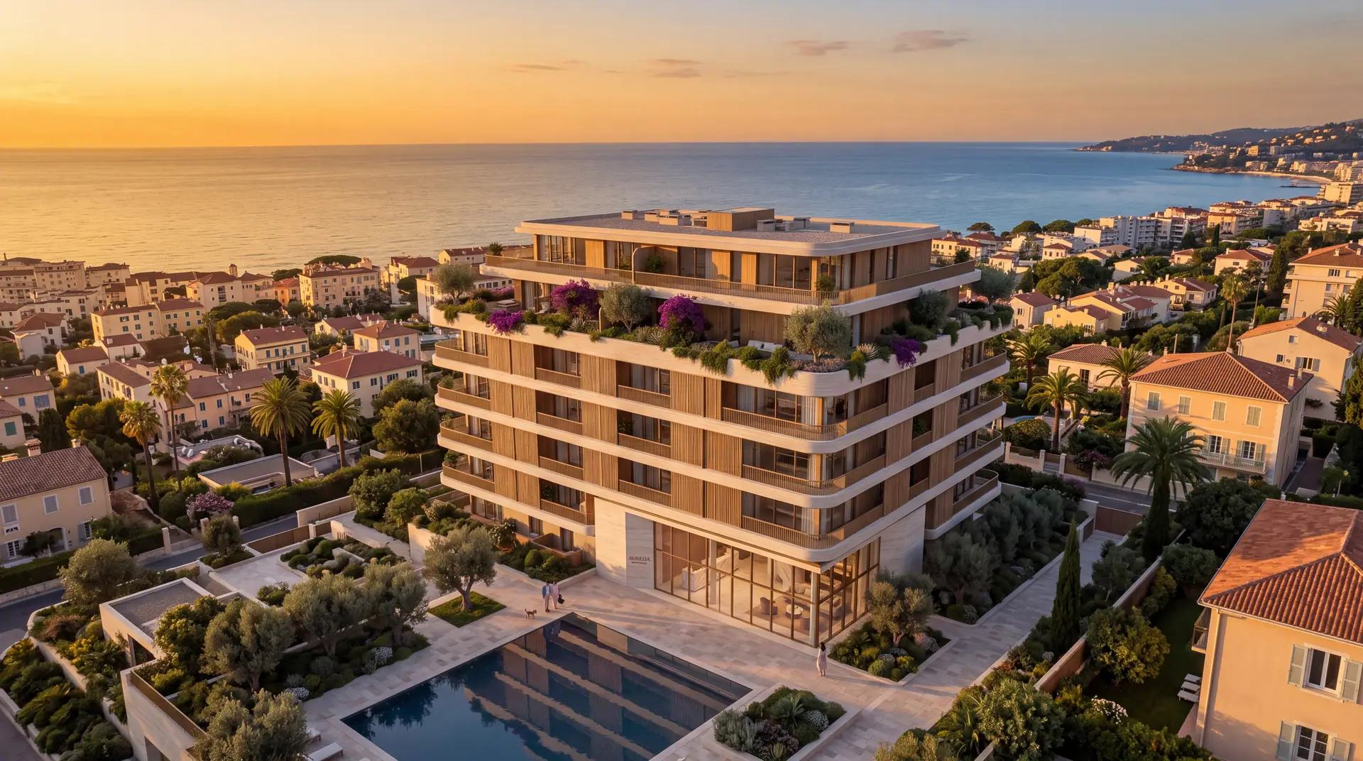 Aerial view of a new apartment building among tiled-roof houses in a coastal town, with a pool, gardens and the sea at sunset